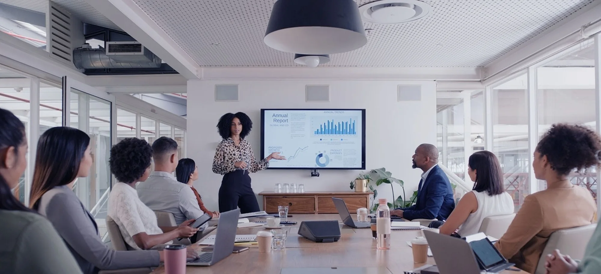 A woman in front of a chart on a TV leading a team meeting in a conference room