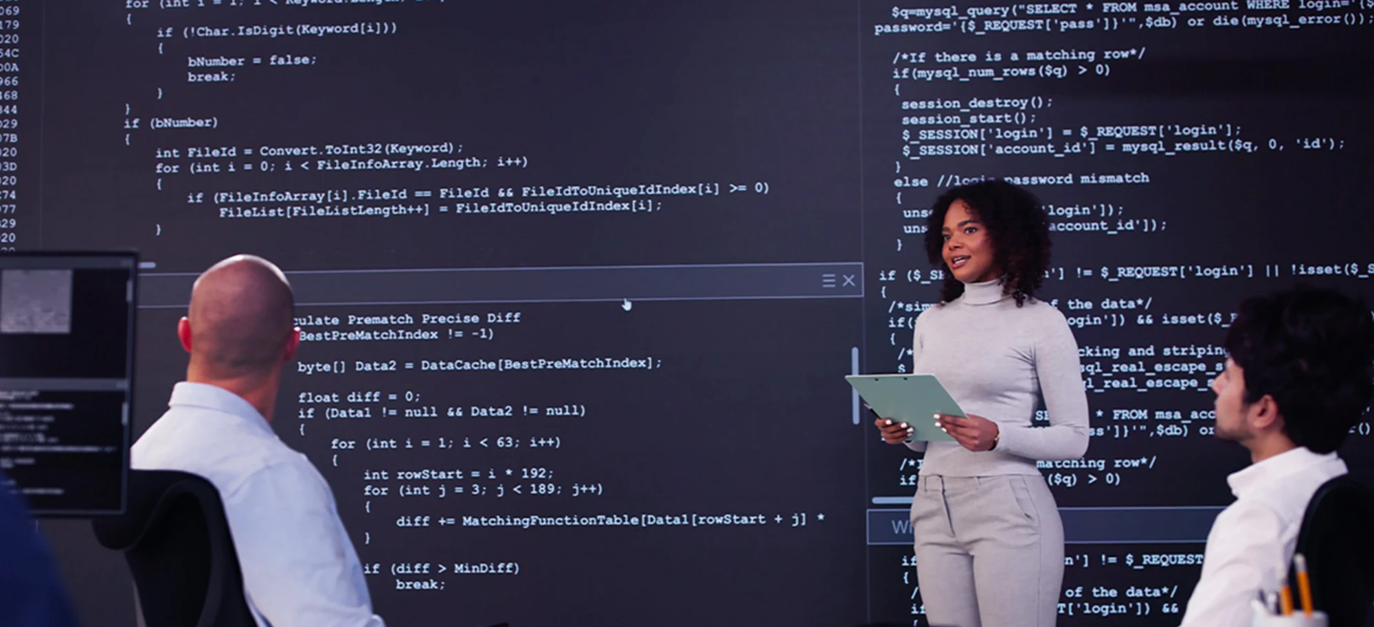 A woman with a clipboard standing in front of a large monitor with code displayed