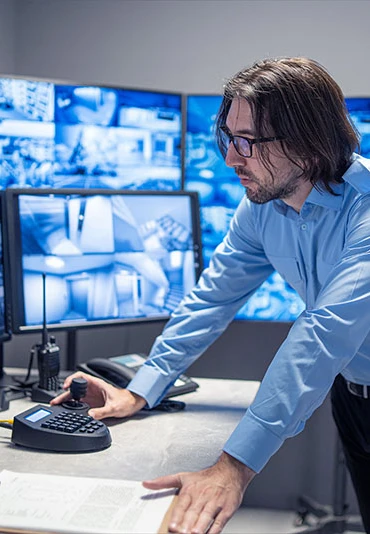 A man using a handheld controller in front of a set of security monitors