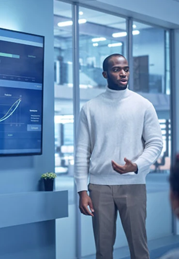 A young man standing next to a monitor addressing a team in a meeting