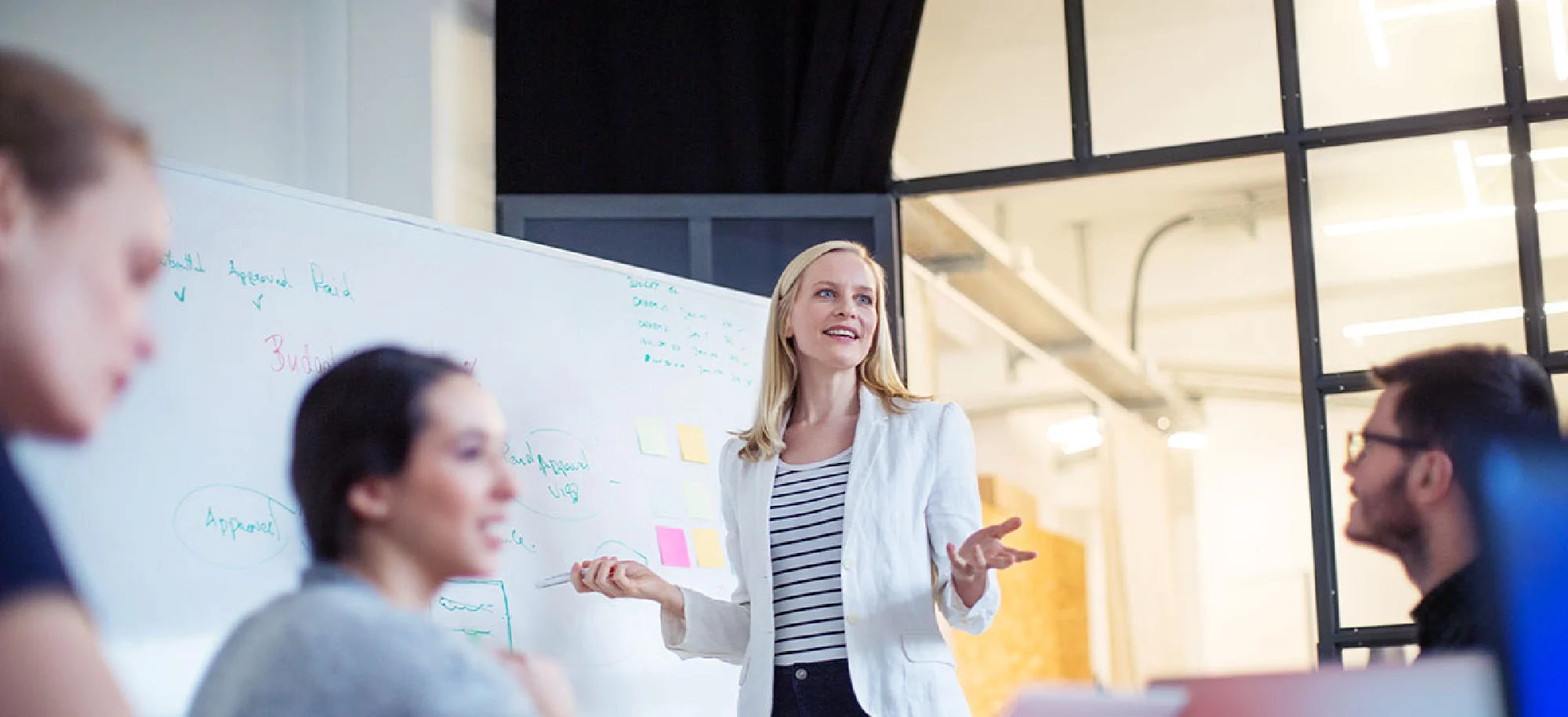 A young female professional standing in front of a white board leading a team meeting